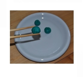 Ein weißer Teller, darauf liegen drei Knetmassekugeln. Eine davon wird gerade mit Stäbchen festgehalten.